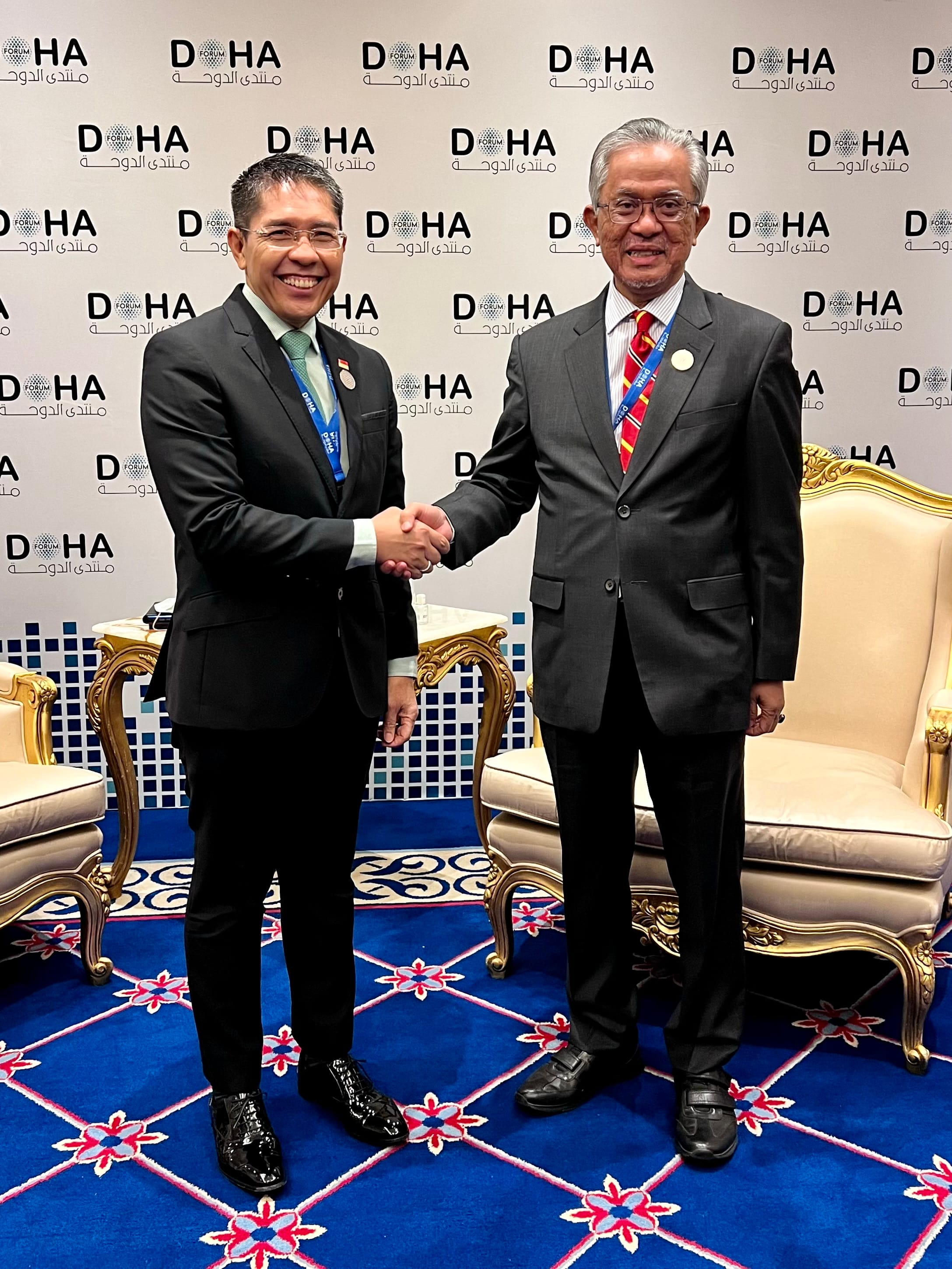 Two men in suits shake hands in front of a Doha Forum backdrop with ornate chairs and a blue patterned carpet.